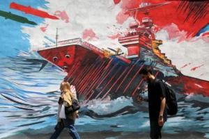 Pedestrians walking past a large outdoor wall mural in Tehran depicting a sinking military warship in rough blue seas under flying missiles, capturing the public atmosphere and ideological messaging directed at the American forward presence.