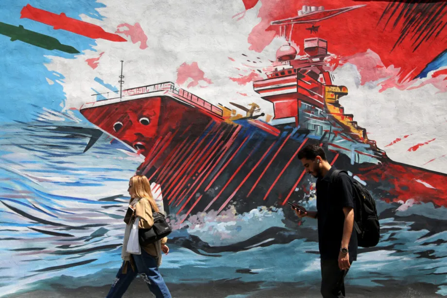نهاية الوجود الأمريكي المتقدم في الخليج الفارسي Pedestrians walking past a large outdoor wall mural in Tehran depicting a sinking military warship in rough blue seas under flying missiles, capturing the public atmosphere and ideological messaging directed at the American forward presence.