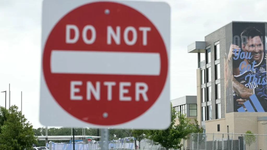 Illegal wars, racism and genocide: USA World Cup 2026 should be boycotted A blurred "DO NOT ENTER" traffic sign stands in the foreground with a building in Kansas City displaying a large promotional football mural in the background, contrasting sports infrastructure with themes of exclusion behind the call to boycott over racism and genocide.