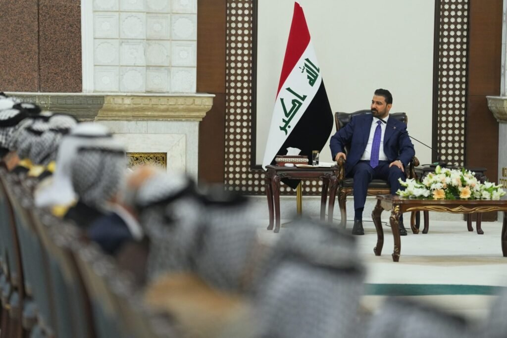 Newly appointed Iraqi Prime Minister Ali al-Zaidi seated in a formal hall next to the Iraqi flag during a meeting, viewed from behind a row of seated officials wearing traditional keffiyeh headdresses, highlighting diplomatic dynamics influencing the US-Iraq relationship.