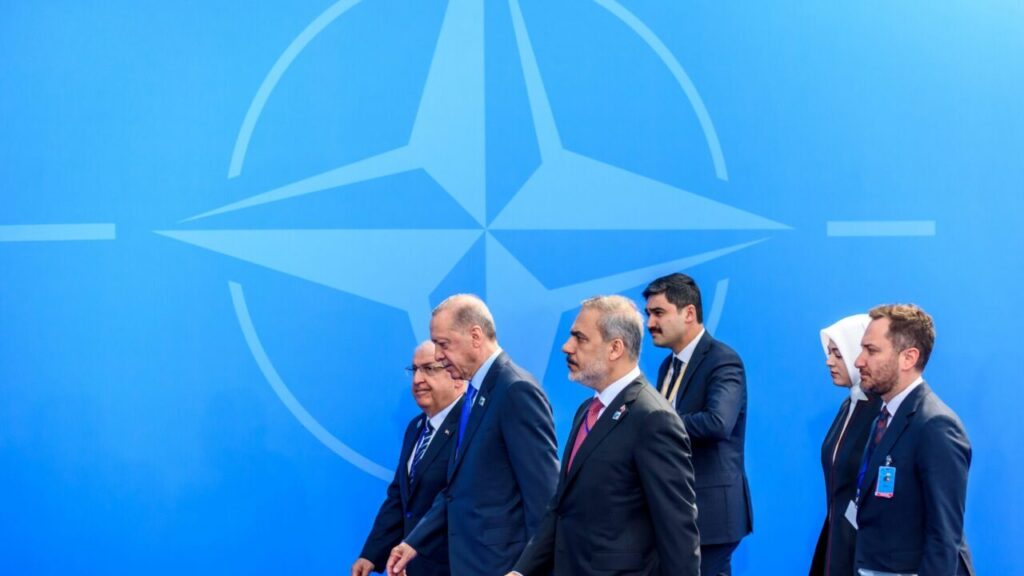 لا تدع تركيا تختطف قمة الناتو Turkish President Recep Tayyip Erdogan walks alongside his diplomatic aides in front of a large blue background featuring the official alliance emblem during a NATO summit.