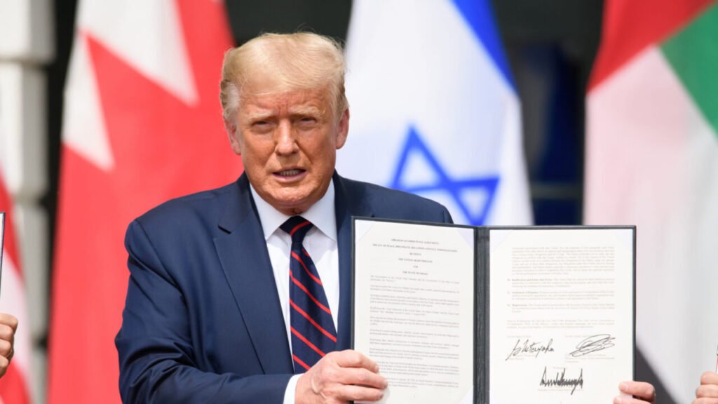 A New US-Led Architecture for the Middle East President Donald Trump stands at a White House podium holding open the signed Abraham Accords document with the flags of Bahrain, Israel, and the UAE blurred in the background, serving as a foundational example of a US-Led Architecture for the Middle East.