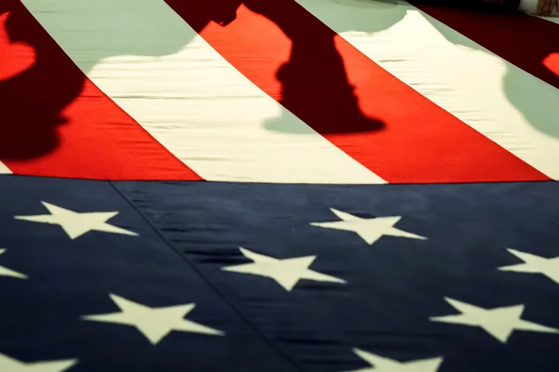 Don’t Give Up on Global Order A close-up view looking down at a large United States flag being held horizontally outdoors, with dark shadows of figures cast across the red and white stripes, visually contrasting the ideals of a unified global order.