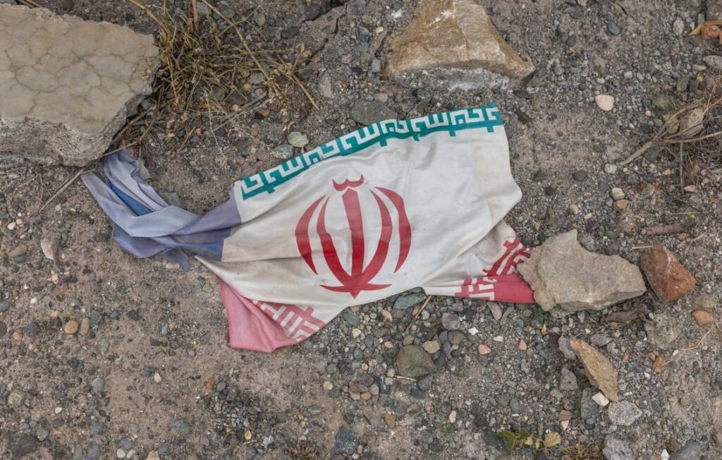 Middle East Conflict Spurs New Displacement A torn and soiled national flag of Iran lying discarded on rough, rocky ground amidst dirt and debris, serving as a symbolic representation of regional instability tied to the Middle East conflict.