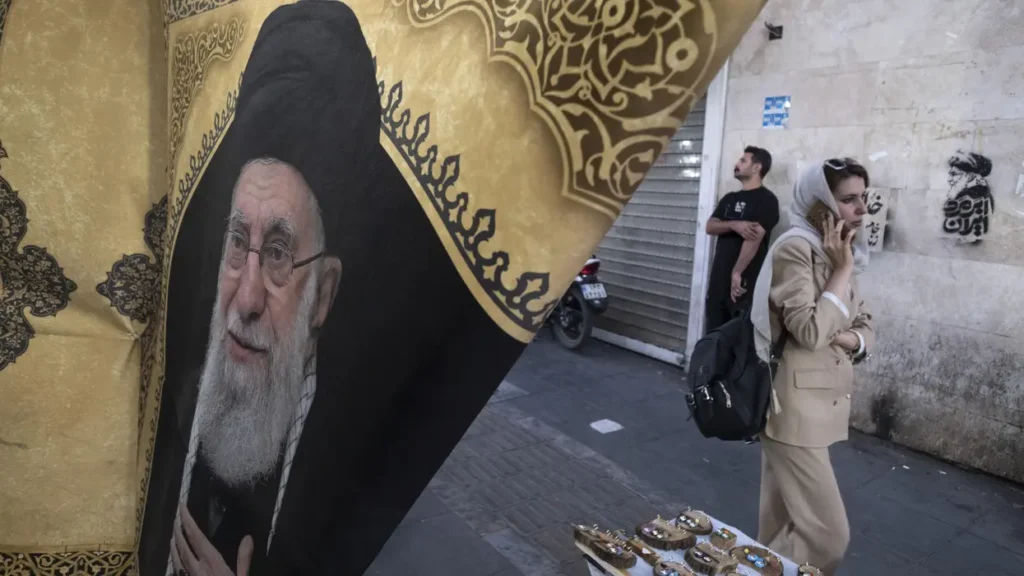 A street scene in Tehran featuring a prominent banner of the late Supreme Leader Ali Khamenei as a woman walks past on her phone, illustrating the domestic landscape following his death in Operation Epic Fury ahead of a potential grand bargain.