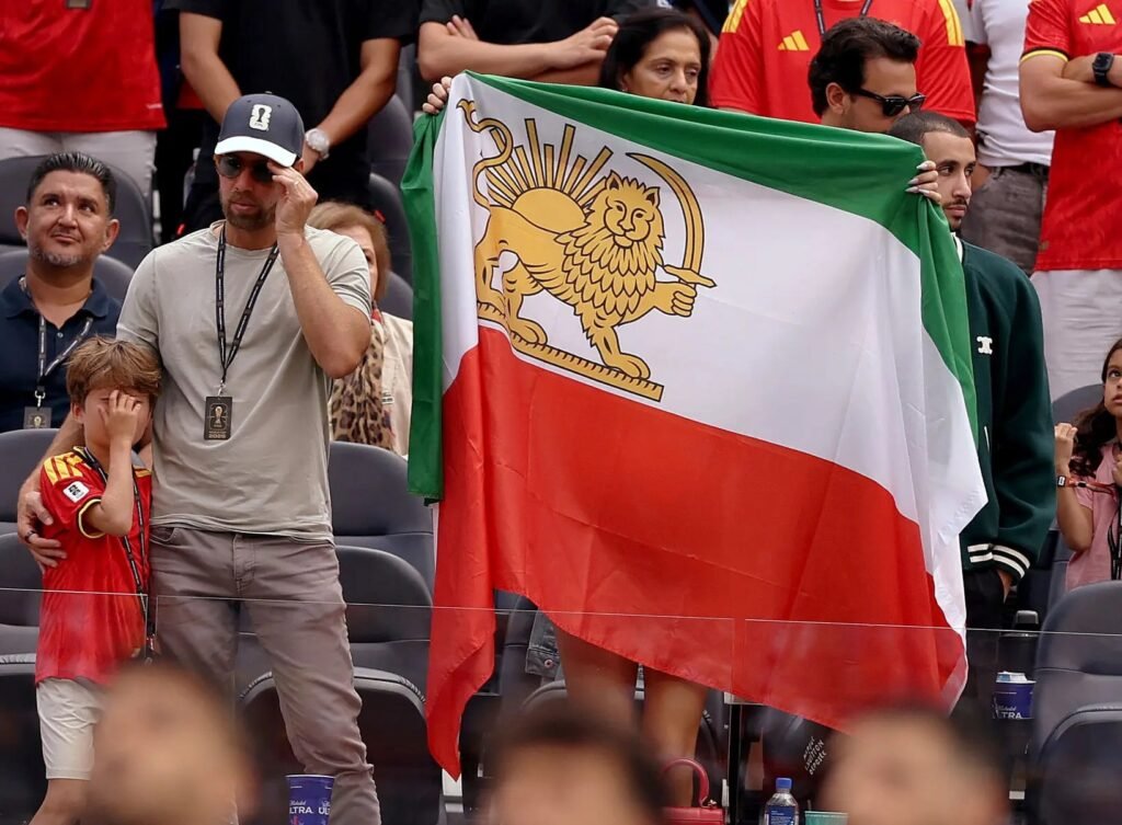 Football fans in stadium stands holding a historical Iranian flag featuring the Lion and Sun emblem, overlooking out-of-focus players on the pitch, illustrating the political complexities surrounding supporting Iran at the World Cup.