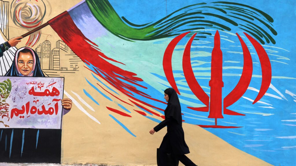 An Iranian woman in a black chador walks past a large street mural featuring political imagery, an Iranian flag emblem stylized as a missile, and a poster of a smiling girl, capturing the ideological climate that will reshape Iran's future.