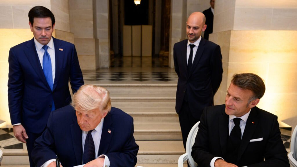 Donald Trump leans forward to sign a document at a table while Emmanuel Macron sits beside him and Marco Rubio stands in the background inside the Palace of Versailles, capturing the high-level diplomatic signing of the US-Iran Memorandum.