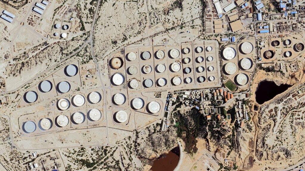 An aerial satellite view shows rows of circular oil storage tanks and industrial infrastructure clustered across the arid terrain of Kharg Island, highlighting the economic vulnerability used as a strategic pressure point.