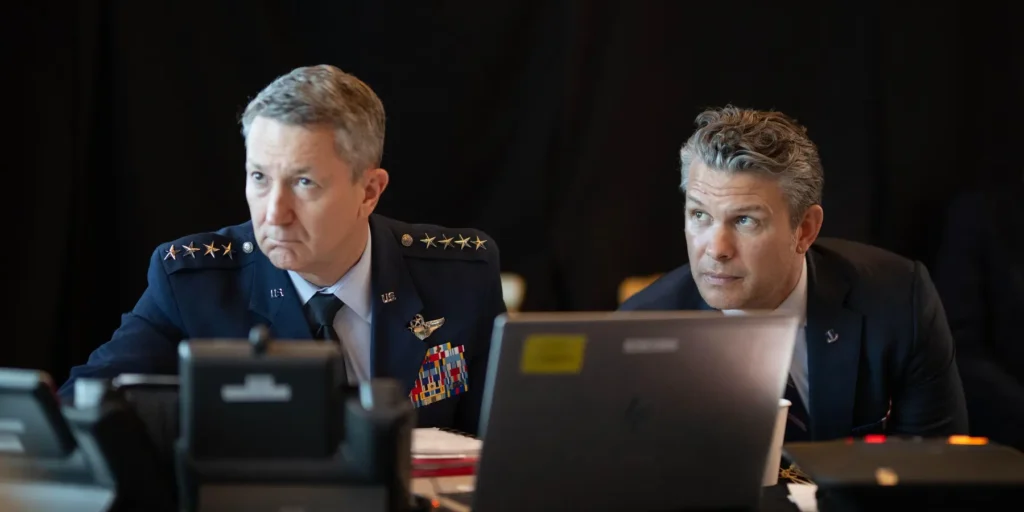 Defense Secretary Pete Hegseth and Joint Chiefs Chairman General Dan Caine seated together behind laptops at a briefing, assessing the military implications of an unpopular war.