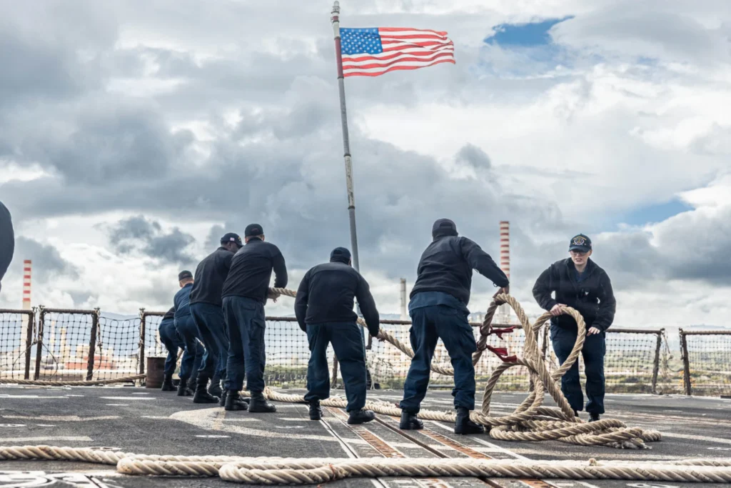 U.S. Navy sailors in dark blue uniforms working together to pull a heavy mooring rope on the deck of a naval vessel under a cloudy sky with the American flag waving, symbolizing the geopolitical shifts and maritime strain that could follow an Iran war change the US role.