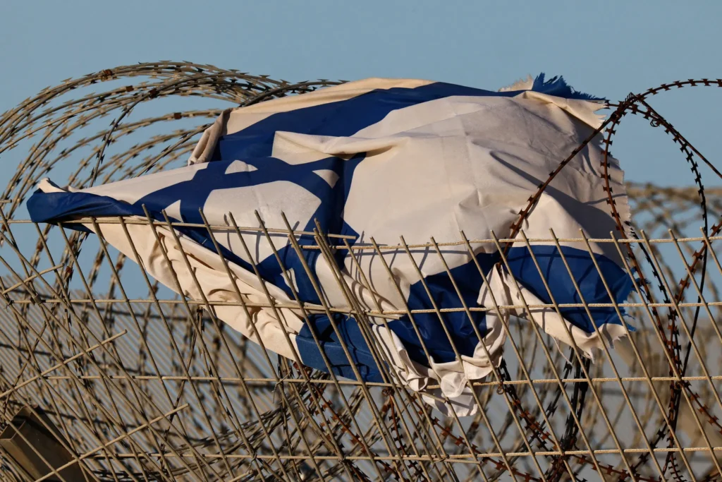 A tattered Israeli flag sits tangled and caught in layers of sharp razor wire atop a metal border fence, symbolically representing the growing isolation facing the Israeli cultural scene.