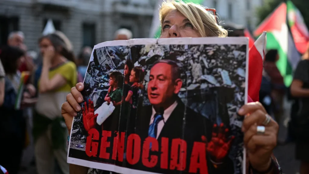 Why Netanyahu wants to wreck Trump’s Iran deal A street demonstrator holds up a protest placard featuring a photo of Israeli Prime Minister Benjamin Netanyahu over a backdrop of urban destruction with the red Spanish/Italian text "GENOCIDA," capturing public outcry against regional geopolitical escalations that threaten diplomatic initiatives like Trump's Iran Deal.