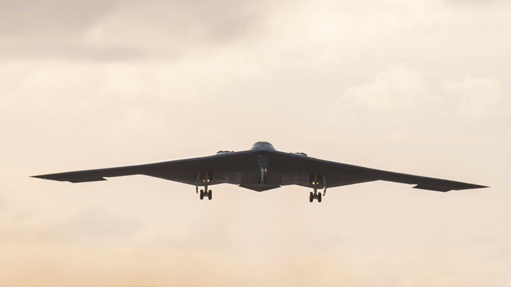 A stealth B-2 Spirit bomber stealth aircraft flying overhead with its landing gear deployed against a hazy, overcast sky, highlighting the military assets deployed to target Iran's nuclear program.