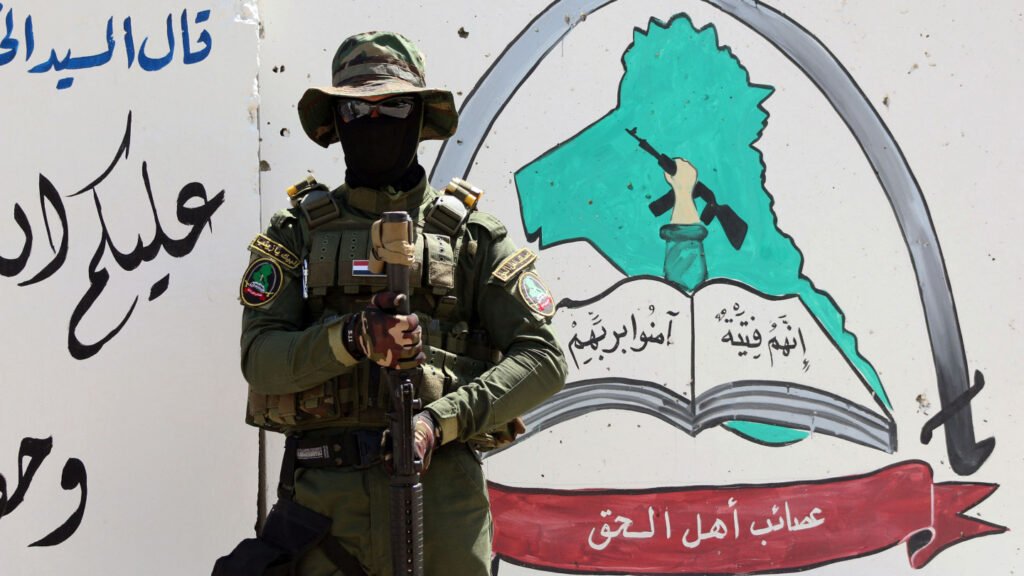 A masked Iraqi paramilitary fighter in full combat gear standing with a rifle in front of a wall mural depicting the official emblem and logo of Asaib Ahl al-Haq.