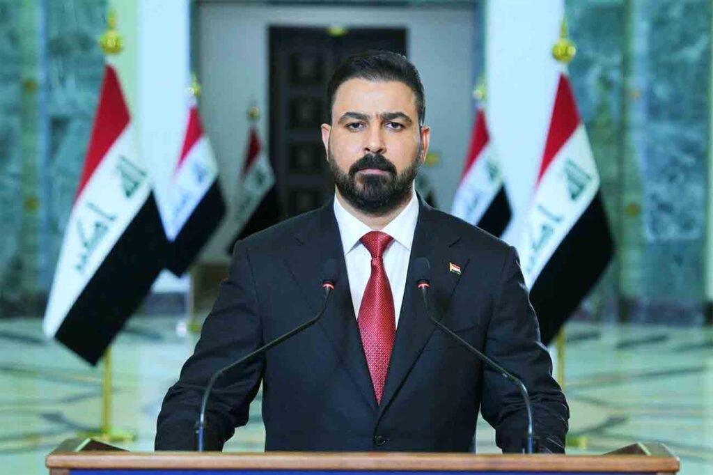 Iraqi Prime Minister Ali al-Zaidi stands formally at a podium wearing a dark suit and red tie with Iraqi flags positioned in the background, framing the institutional setting behind the debate on the state's monopoly on arms.