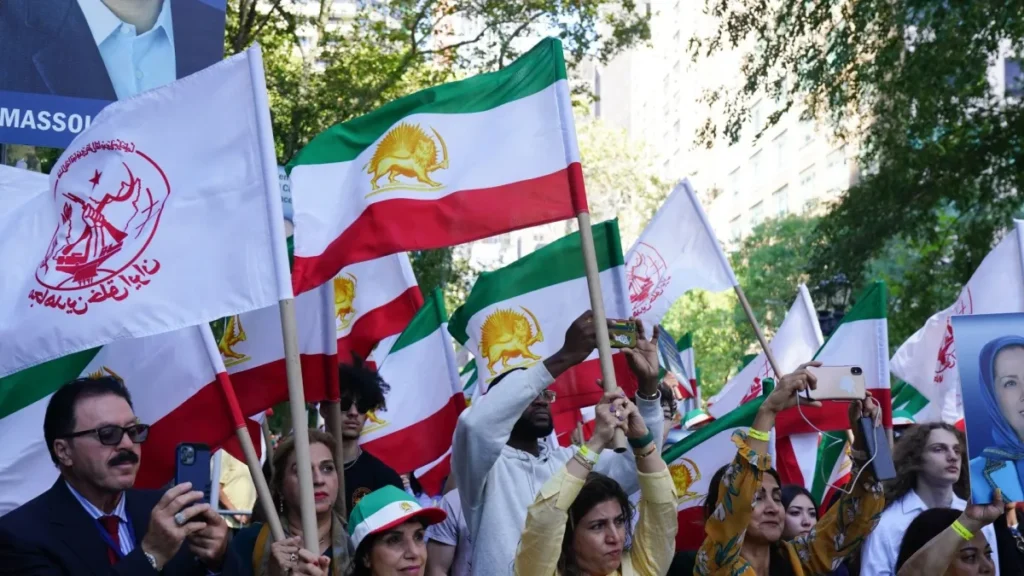A crowd of demonstrators gathered outdoors waving Iranian Lion and Sun flags used by monarchist supporters of Reza Pahlavi alongside white flags with the logo of the People's Mojahedin Organization of Iran (MEK), while holding up smartphones and political portraits during a protest, capturing the political mobilization associated with Iran's opposition.