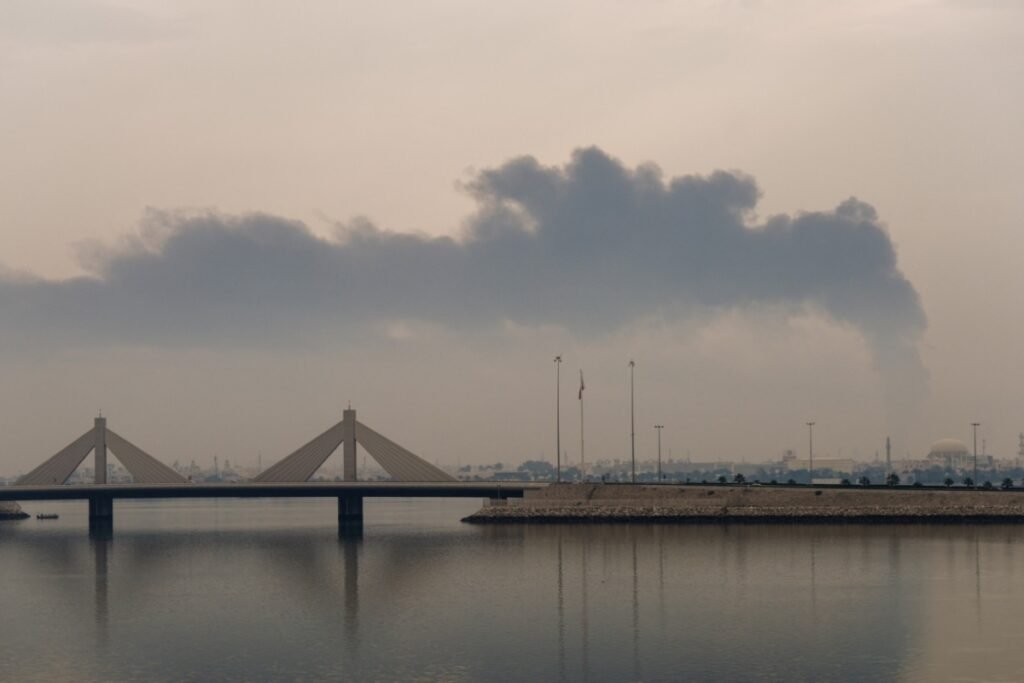 Thick dark smoke rises into a hazy sky above a coastal skyline beyond a modern cable-stayed bridge spanning the water, visually representing the security vulnerabilities that put Bahrain in Iran's crosshairs.