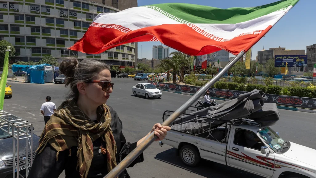 A woman wearing sunglasses and a patterned scarf holds a large Iranian flag waving in the wind above a bustling Tehran city street with cars and buildings, capturing the national domestic landscape behind the narrative that Iran Won the War.