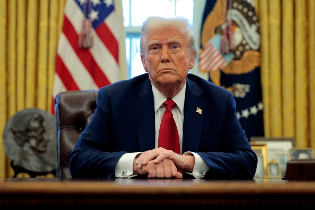 Donald Trump sits at a desk in the Oval Office with his hands clasped, flanked by the American flag and the presidential seal, providing a formal backdrop to discussions on whether an MOU qualifies as a lasting peace deal.