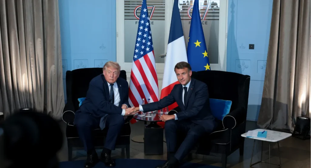 Taking the Pulse: Is European Diplomacy on Iran Outdated? US President Donald Trump and French President Emmanuel Macron shake hands while seated in black armchairs in front of the US, French, and European Union flags, visually capturing the strategic alignment and debates surrounding European diplomacy on Iran.