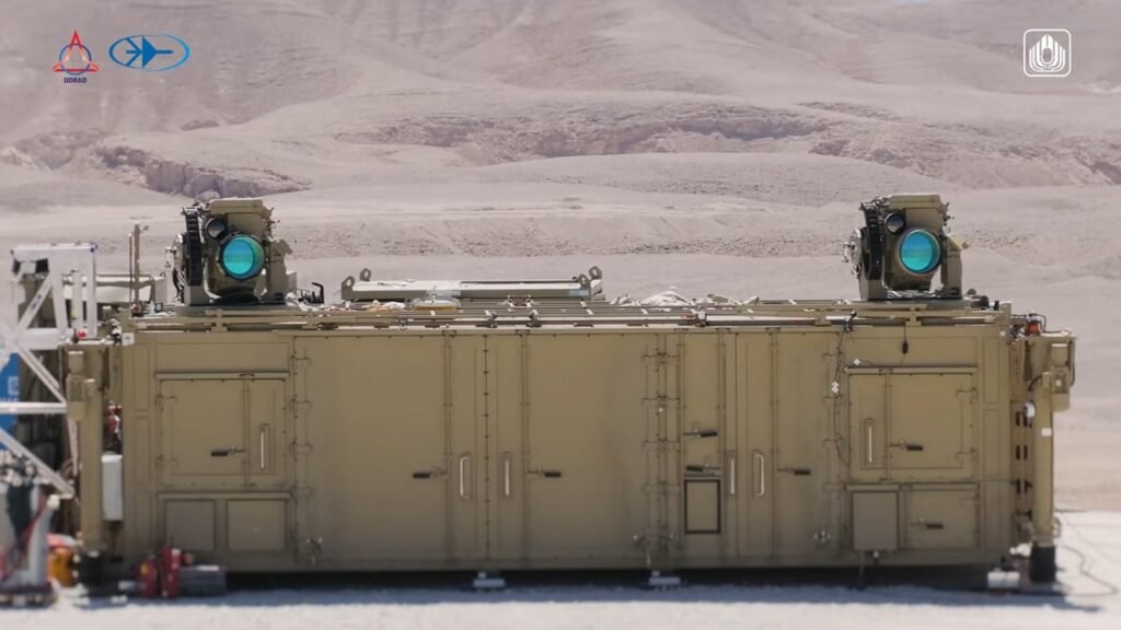 A large, military-tan container platform sits in a desert environment equipped with two elevated Iron Beam air defense batteries featuring large blue glass lenses, demonstrating the tactical infrastructure behind Israel's new laser weapon.