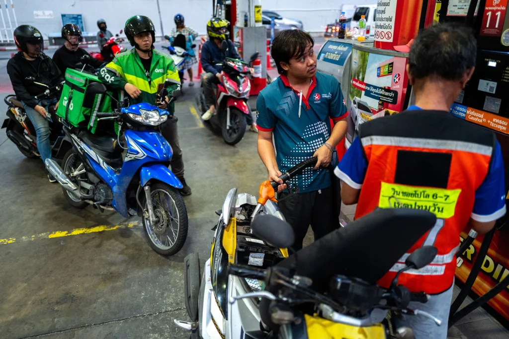 Motorcyclists queuing at a busy gas station in Bangkok while a station attendant pumps fuel, highlighting the vulnerabilities of Asia's critical energy dependence during geopolitical crises.