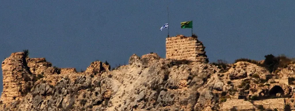 A wide view of the ancient stone ruins of Beaufort Castle perched on a rocky hilltop, featuring the Israeli flag and the green flag of Hezbollah flying side by side from the highest tower, symbolizing the strategic border contestation and Pressure on Hezbollah.