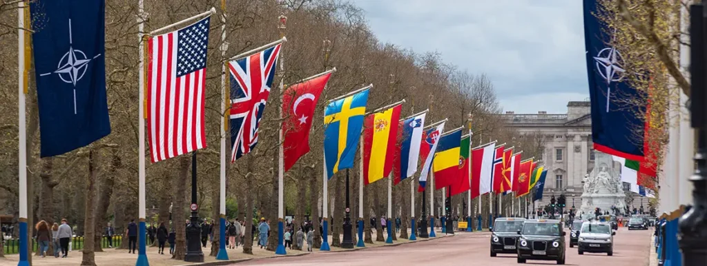 A wide street view of The Mall in London lined with international flags including the US, UK, Turkey, and NATO banners as vehicles drive past, representing the collective security framework brought into focus by The Iran Conflict.