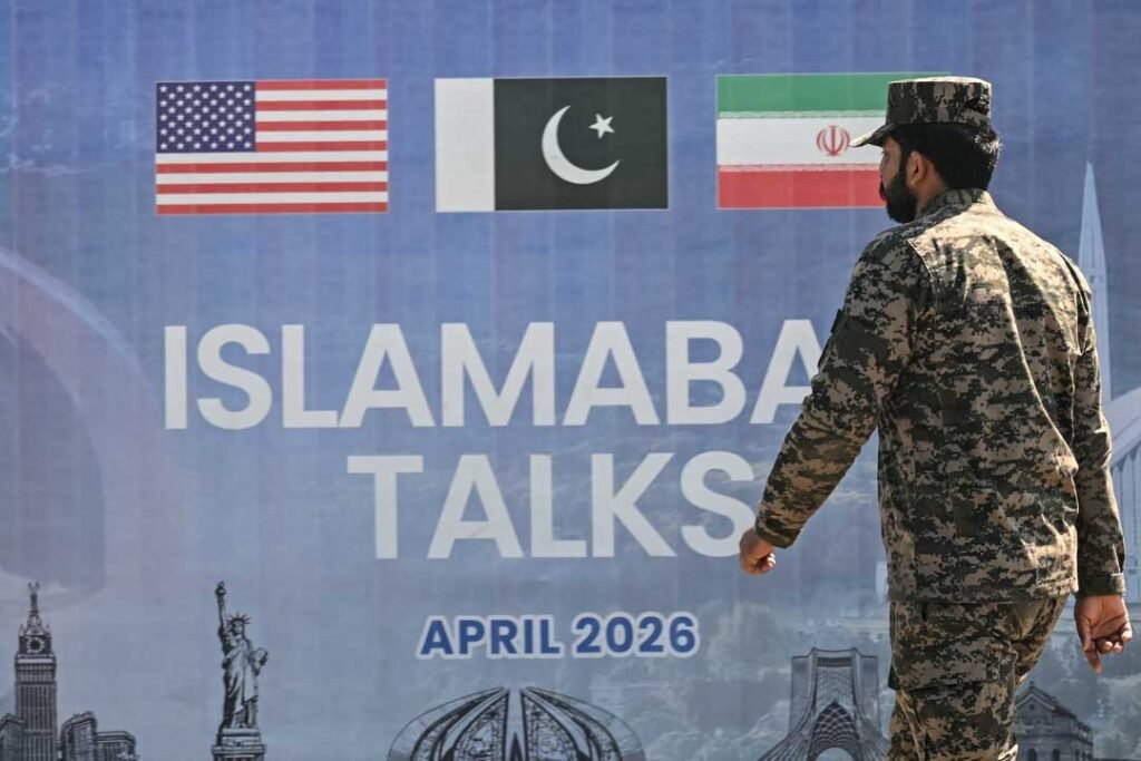 A soldier in camouflage uniform walks past a large summit backdrop reading "ISLAMABAD TALKS APRIL 2026" featuring the flags of the United States, Pakistan, and Iran, visually representing diplomatic mediation efforts aimed at a U.S.-Iran breakthrough.