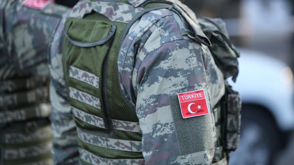 A close-up view from behind of a soldier wearing camouflage tactical gear and a vest with a prominent red Turkish flag patch reading "TÜRKIYE" on the sleeve, symbolizing the regional defense postures involved in reshaping Turkey.