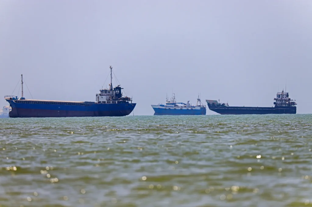 Four Ways the Gulf Makes Iran’s Hormuz Toll Backfire Several large cargo ships and industrial maritime vessels floating on choppy green seawater under a hazy blue sky, illustrating the commercial shipping dynamics impacted by the proposed Hormuz toll.