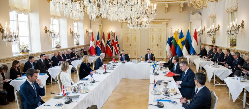 How War in Iran is Impacting the Baltic States A large diplomatic summit meeting features delegates seated around a U-shaped white table in an ornate, classical hall lined with Nordic, Baltic, and European flags, highlighting regional strategic discussions influenced by the ongoing war in Iran.