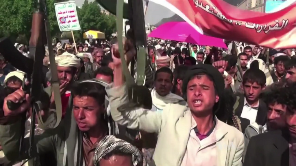 A large crowd of Houthi supporters marches through a street with some individuals chanting and holding up rifles next to banners, illustrating the mobilization and regional alignment of the Houthis.