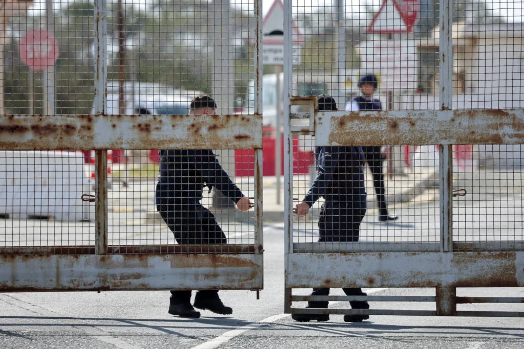 Two security guards in dark uniforms pull shut a large, rusted metal mesh security gate across a roadway, capturing the physical barriers and shifting strategies that define the modern limits of the American way of war.