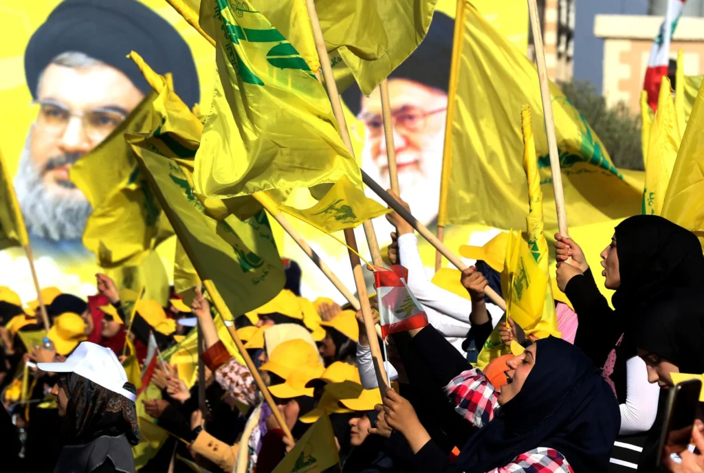 A crowd of supporters waving bright yellow Hezbollah flags and Lebanese flags during a public rally with massive background portraits of regional leaders, highlighting the ideological alignment and geopolitical Protection of Hezbollah.