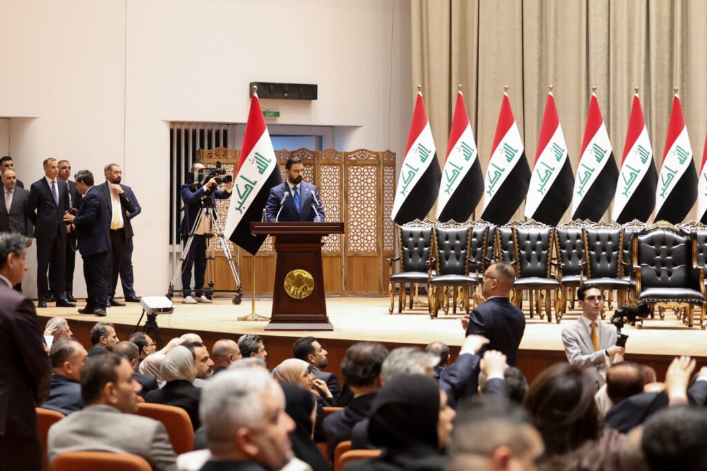 عراق در میان آشفتگیهای منطقهای دولت جدیدی تشکیل میدهد Iraq's new Prime Minister Ali al-Zaidi delivering a speech from a wooden podium at a parliamentary session with multiple national flags and black leather chairs lined up on stage, marking the legislative procedures as Iraq Forms New Government.