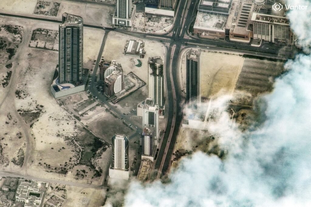 خلیج فارس و ایران: دو مدل که به نظر میرسد غیرقابل سازش هستند An aerial satellite view shows a cluster of modern high-rise glass skyscrapers standing next to undeveloped desert terrain near a coast shrouded in clouds, symbolizing the stark structural contrasts underlying the relationship between The Gulf and Iran.