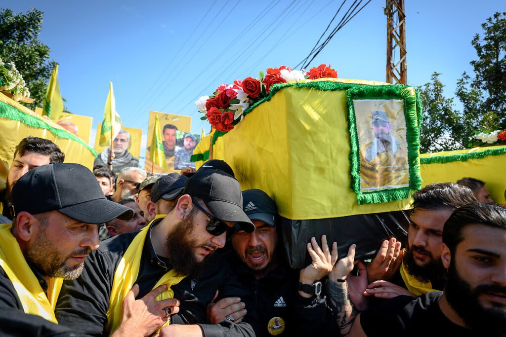 After massive Israeli attacks, how is Hezbollah still fighting? A crowd of men wearing black shirts and caps carry a coffin wrapped in a yellow flag adorned with a portrait during an outdoor funeral procession, providing visual context to the socio-political environment and the community dynamics behind why Hezbollah still fighting.