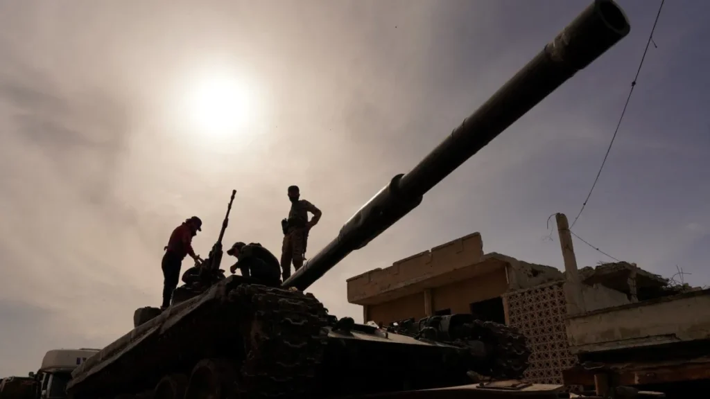 A low-angle silhouette shot shows Syrian soldiers standing on a tank with its prominent gun barrel pointing upward against a bright, hazy sky near a damaged concrete structure, establishing visual context for the regional geopolitical maneuvers surrounding Syria’s Sharaa.