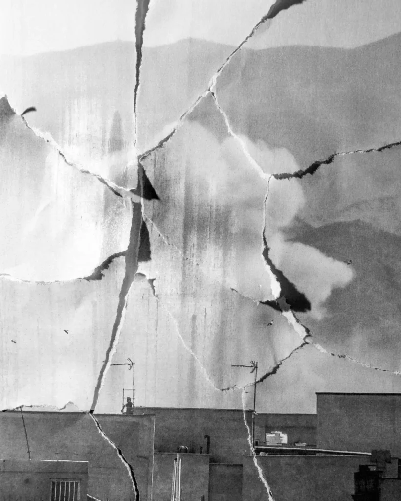 A black-and-white image of a cityscape featuring buildings and rooftops appears heavily fractured and torn into shattered pieces, symbolically representing the widespread devastation and ruin caused by the Middle East war.