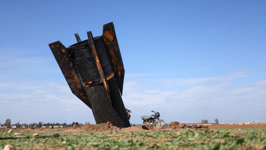 The charred tail section of a large ballistic missile embedded in the ground in an open field next to a motorcycle, illustrating the remnants of conflict affecting Syria after the Iran war.