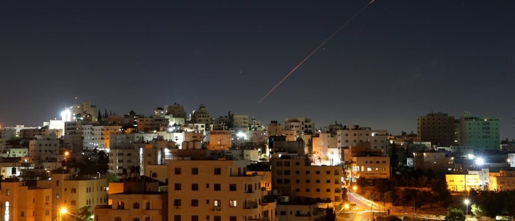 A wide night panoramic view of an urban skyline under a dark sky lit up by a sharp, diagonal red light streak from a missile trajectory, capturing the volatile military background that threatens whether Iran negotiations can endure.