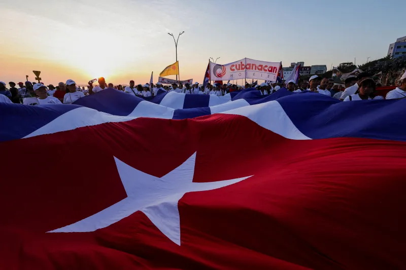 روز بعد در کوبا A massive Cuban flag featuring its prominent white star on a red triangle is carried horizontally by a large crowd marching outdoors at sunrise, highlighting the deep-seated political mobilization and nationalist sentiments often central to debates surrounding a potential Cuba Regime Change.