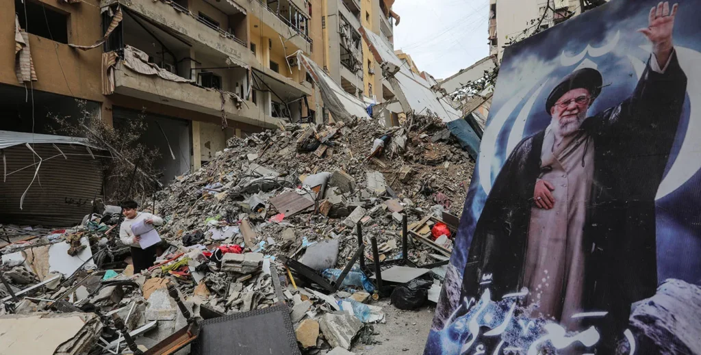 A large billboard featuring an image of the late Iranian leader stands prominently beside a massive pile of concrete rubble and heavily damaged apartment buildings in Lebanon, highlighting the destructive aftermath of regional conflicts involving Hezbollah.