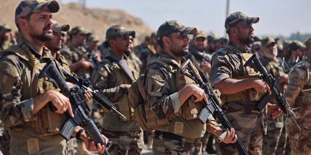 A line of uniformed Saraya al-Salam paramilitary fighters stands in formation holding assault rifles during a ceremony in Samarra, visualizing the political and security challenges surrounding Iraq's armed factions.