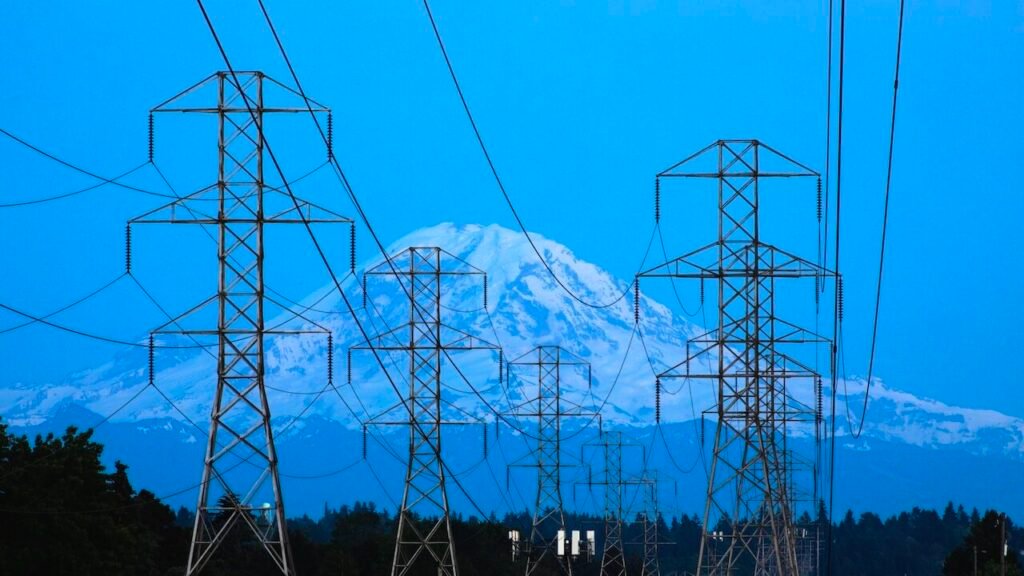 چرا زیرساختهای حیاتی آمریکا میتواند جذابترین هدف ایران باشد High-voltage electrical power lines and transmission towers stretch across the landscape with the snow-covered peak of Mount Rainier in the background, highlighting the domestic systems vulnerable as a targeted critical infrastructure.