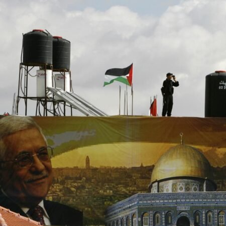 A security officer standing on a rooftop with a Palestinian flag and water tanks, positioned above a large banner featuring Mahmoud Abbas and the Dome of the Rock, capturing the political environment as Fatah Meets in Ramallah.