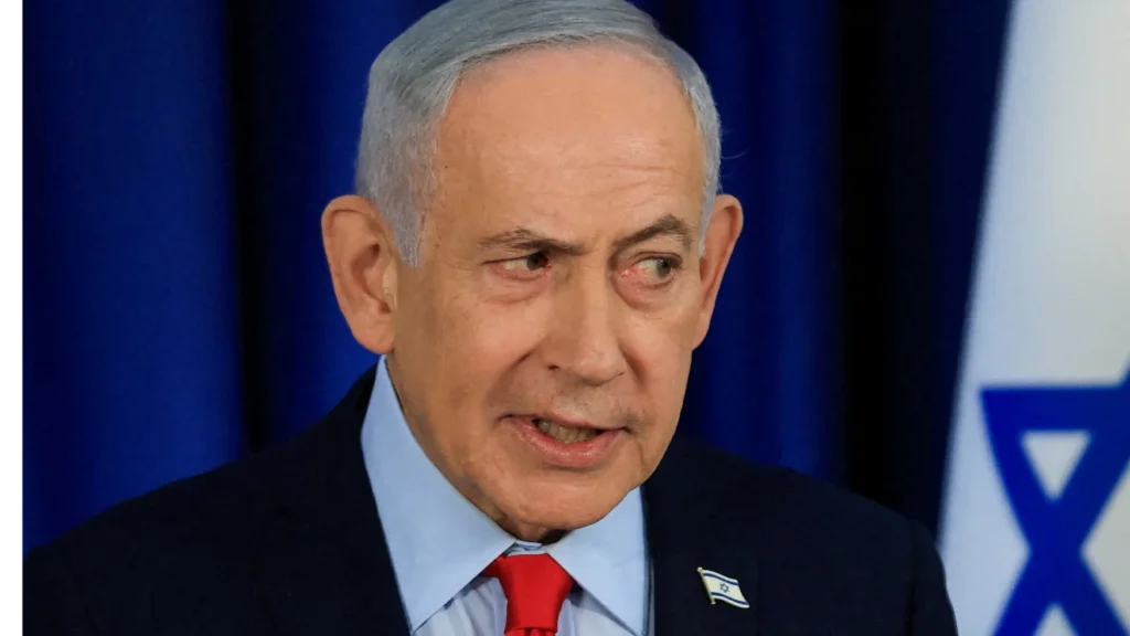 A close-up portrait of Israeli Prime Minister Benjamin Netanyahu wearing a dark suit, light blue shirt, and red tie while speaking, with a blue backdrop and a partial view of the Israeli flag behind him, capturing the political tensions underlying the assertion that Netanyahu won't stop.