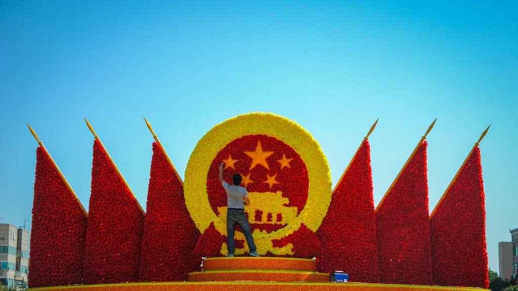 The Next Front in US-China Competition Is the Homefront A worker stands on a stage arranging a massive floral installation of the national emblem of the People’s Republic of China flanked by red floral flags, capturing the soft-power initiatives underlying the US-China Competition.